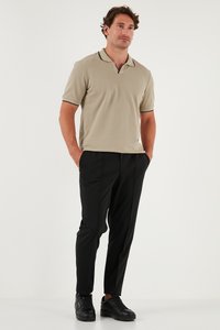 Mees seisab, käed taskus, seljas beež lühikeste varrukatega polo, mustad püksid ja mustad tossud valgel taustal.