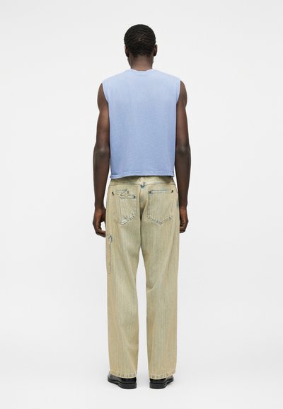 Άτομο που στέκεται εμφανίζοντας την πλάτη, φοράει ανοιχτό μπλε αμάνικο τοπ, ανοιχτό μπεζ παντελόνι με φαρδιά πόδια και μαύρα παπούτσια σε απλό φόντο.