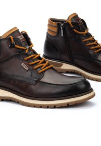 Bottes en cuir marron avec des accents beiges, dotées de lacets orange, d'une fermeture éclair sur le côté et d'une semelle en caoutchouc texturée. Des détails cousus sont présents.