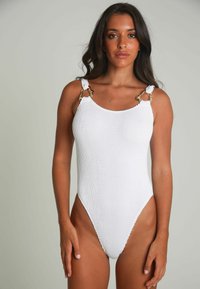Maillot de bain une pièce blanc en tissu texturé, avec des éléments en or sur les bretelles nouées et des jambes hautes, créant une silhouette élégante.