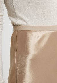 Sukienka A-line z satyny w jasnym beżu, o gładkiej fakturze, z prążkowanym paskiem w talii. Materiał ma lekki połysk i subtelne drapowanie.