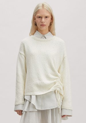 Vrouw met lang blond haar draagt een oversized crème gebreide trui over een gestreepte hemdjurk, staand tegen een effen grijze achtergrond.