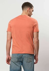 Homme vu de dos, portant un t-shirt pêche uni et un jean bleu clair sur un fond uni gris clair.