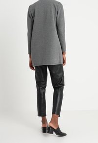 Henkilö, jolla on päällään pitkä harmaa teksturoitu neule, mustat nahkahousut ja mustat korkeat liukupohjaiset kengät, seisoo valkoista taustaa vasten.