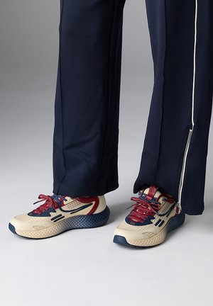 Person trägt marineblaue Hose mit seitlichen Reißverschlüssen und beige Sneakers mit roten und marineblauen Akzenten, steht auf einem hellgrauen Boden.