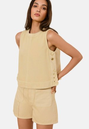 Woman wearing sleeveless beige top with side buttons and matching beige shorts with front pockets, posing with hands behind her back.