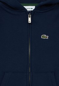 Navy zip-up hoodie made of cotton, featuring a green-lined hood, front pockets, and a small embroidered crocodile logo on the chest.