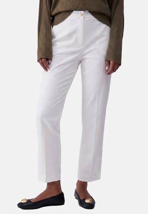 Pantaloni bianchi a gamba dritta con bottone frontale, abbinati a ballerine nere con grande decorazione dorata, indossati con un maglione marrone a coste.