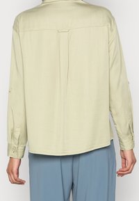 Chemise à manches longues vert clair présentant une coupe droite, des détails de couture à l'arrière et des poignets boutonnés. Fabriquée dans un tissu lisse et léger.