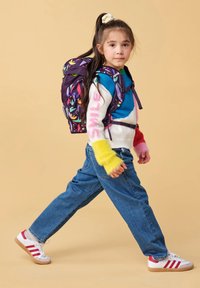 Jong meisje loopt met een kleurrijke rugzak, draagt een multicolor trui, blauwe jeans en witte sneakers met rode strepen.