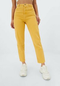 Gula högmidjade jeans i denim, med smal passform, fem fickor och knappstängning, kombinerade med vita sneakers.