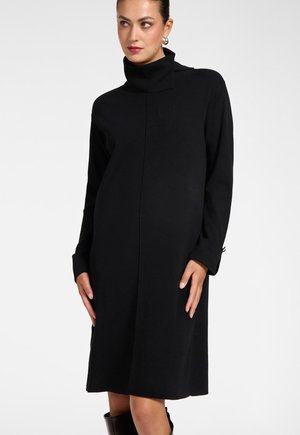 Vrouw draagt een zwarte jurk met lange mouwen en coltrui, staat met haar handen bij haar heupen en kijkt lichtjes opzij.
