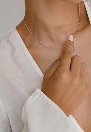 Silver necklace with a delicate, curved design, worn around the neck. Hand with a neutral nail polish gently touches the necklace.