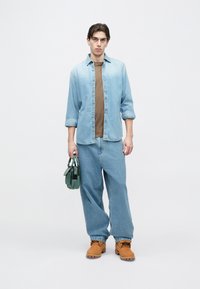 Camisa de mezclilla azul claro, camiseta color canela, pantalones anchos de mezclilla azul claro, botas naranjas, sosteniendo una bolsa verde de forma rectangular con herrajes.