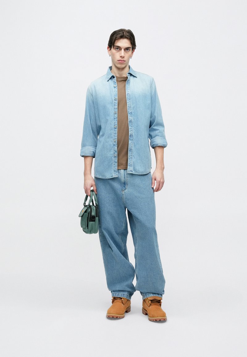 Camisa de mezclilla azul claro, camiseta color canela, pantalones anchos de mezclilla azul claro, botas naranjas, sosteniendo una bolsa verde de forma rectangular con herrajes.