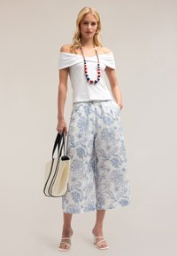 Top blanco off-the-shoulder, pantalones anchos con estampado floral en azul y blanco, sandalias de tacón blancas y un bolso beige con acentos oscuros.