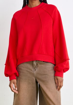 Personne portant un sweat-shirt rouge vif à manches longues avec des manches bouffantes et un pantalon large beige sur fond blanc.