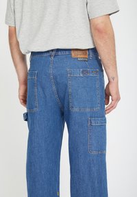 Los pantalones cargo de mezclilla tienen un corte holgado, múltiples bolsillos y una cinturilla alta. El color es un azul medio, con una tela texturizada y duradera.