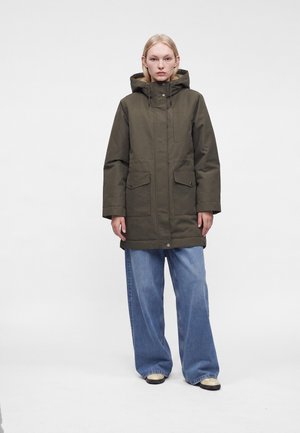 Jeune personne debout, mains détendues, portant un manteau à capuche vert olive, un jean bleu ample et des chaussures beiges sur un fond blanc uni.