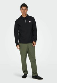 Schwarzer Reißverschluss-Pullover mit Kragen, versehen mit einem kleinen Logo-Patch. Getragen mit grünen Hosen und schwarzen Sneakers. Glatte Textur, lässige Passform.