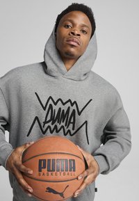 Sweat à capuche gris avec un logo noir "PUMA" aux accents déchiquetés. Le modèle tient un ballon de basket avec une étiquette noire "PUMA BASKETBALL" en évidence.