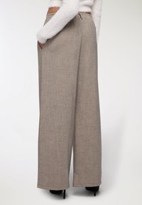 Donna che indossa pantaloni beige a vita alta e gamba larga con tasche laterali e scarpe con tacco nero, abbinati a un top bianco peloso a maniche lunghe.