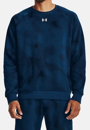 Mand iført mørkeblå tekstureret langærmet sweatshirt og matchende bukser med et hvidt Under Armour-logo på brystet.