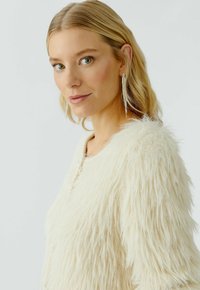 Blonde vrouw met groene ogen draagt lange zilveren oorbellen en een crèmekleurige, fluffy textuur-jacket, terwijl ze zacht naar de camera kijkt.