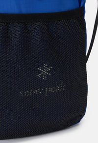 Sac bleu avec poche en maille noire, portant un logo. Le design comprend un tissu lisse et une maille texturée pour la respirabilité.