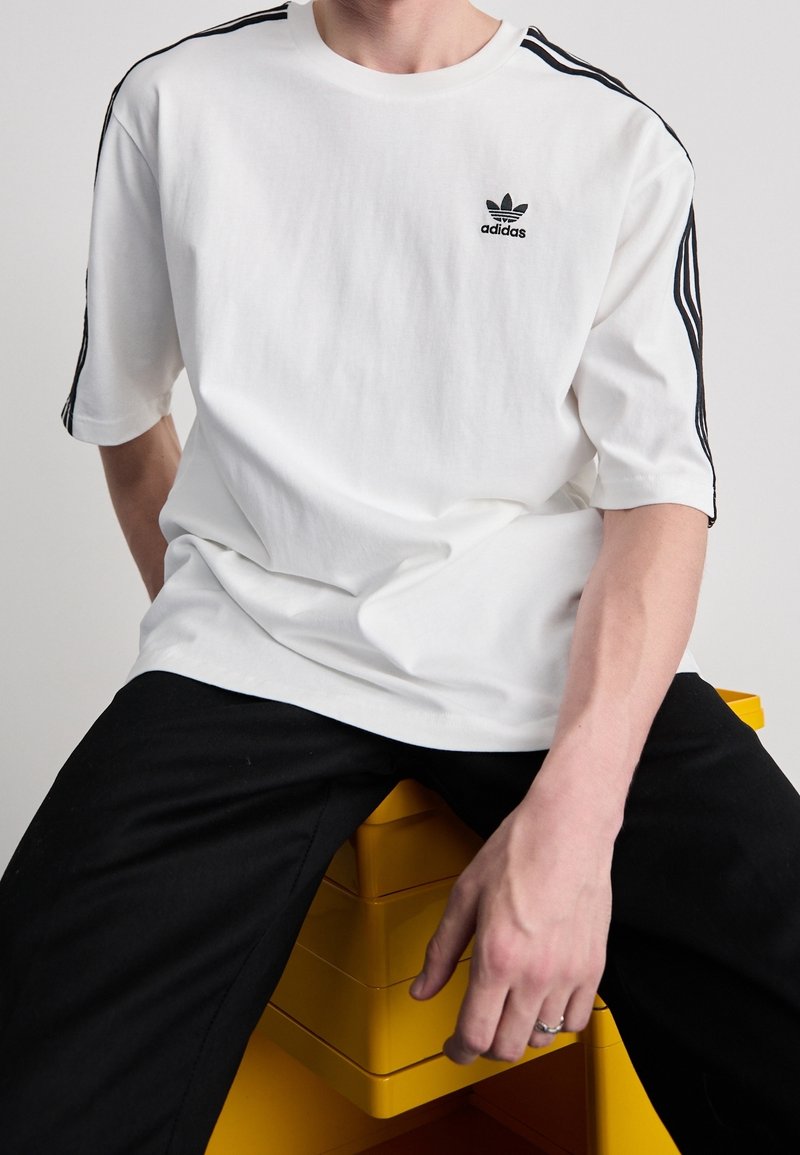 Bílý bavlněný tričko s krátkým rukávem, s černým logem Adidas a třemi černými pruhy na každém rameni. Nosit s černými kalhotami.