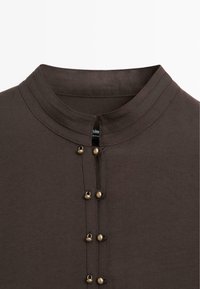Donkerbruine top met een hoge halslijn, voorzien van een rij decoratieve knopen in messingstijl langs de voorlijst en subtiele stiksel details.