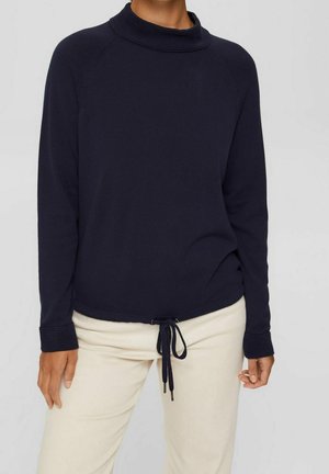 Osoba ubrana w granatowy sweter z długim rękawem z prążkowanymi mankietami i dekoltem, w połączeniu z jasnobeżowymi spodniami z troczkiem z przodu.