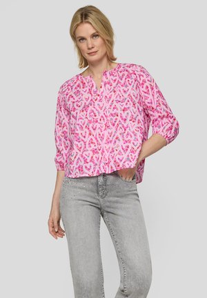 Blonde Frau trägt eine pinke Bluse mit Herzmuster und dreiviertel Ärmeln, kombiniert mit hellgrauen Jeans mit Strassdetails.