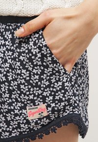 Námořnické šortky s bílým květinovým vzorem, se krajkovým lemem na spodním okraji a štítkem, na kterém je napsáno "Sunshine SuperDry." Viditelné kapsy.