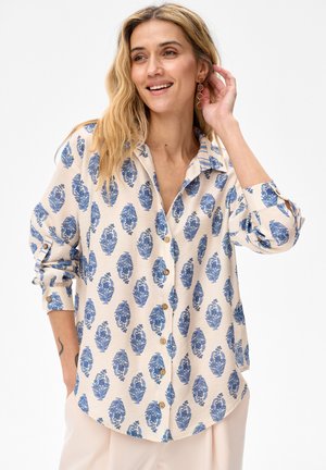 Uśmiechnięta kobieta o falowanych blond włosach nosi beżową koszulę na guziki z niebieskim kwiatowym wzorem, z długimi rękawami podwiniętymi oraz jasne spodnie.