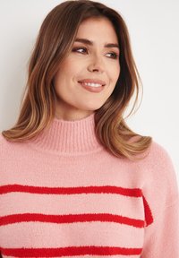 Maglione rosa con un alto colletto a costine, caratterizzato da strisce orizzontali rosse. La texture è morbida e lavorata a maglia, con un design a vestibilità rilassata.