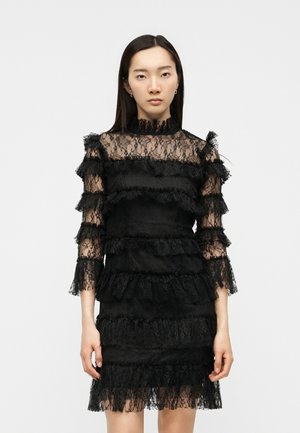 Γυναίκα με ίσια σκούρα μαλλιά που φορά μαύρο δαντελένιο φόρεμα με βολάν και μανίκια μέχρι τον αγκώνα, στέκεται μπροστά σε ένα απλό λευκό φόντο.