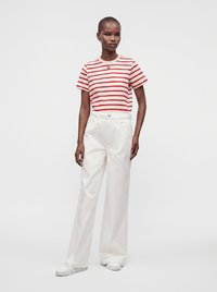Femme portant un t-shirt à manches courtes rayé rouge et blanc, un pantalon blanc taille haute à jambes larges et des baskets blanches, debout devant un fond uni.