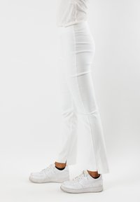 Pantaloni bianchi aderenti con una texture liscia e orlo svasato, abbinati a sneaker bianche. Nessun motivo visibile o dettagli in metallo.