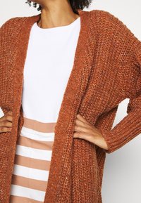 Rustkleurige gebreide cardigan met een open voorkant, geribde mouwen en een textuurafwerking. Gelaagd over een witte top en een gestreepte rok.
