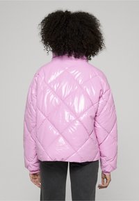 Różowa, błyszcząca kurtka puchowa z pikowanym wzorem w diamenty, wysokim kołnierzem i luźnym krojem. Noszona na szarych spodniach, eksponująca swoją lekką teksturę.