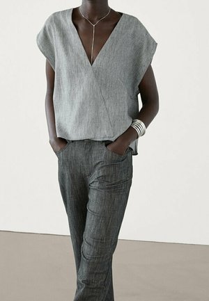 Persona che indossa una maglia grigia stile incrociato a maniche corte, pantaloni grigio scuro, collana d'argento e braccialetti d'argento a strati con le mani nelle tasche.