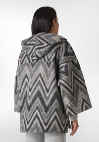 Fleecejakke med hætte, grå og sort zigzag-mønster, løs pasform og brede ærmer. Den teksturerede stof skaber et hyggeligt udseende.