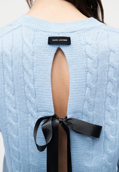 Pull bleu clair en maille torsadée avec une ouverture dos en forme de goutte maintenue par un ruban en satin noir et une étiquette Marc Jacobs au niveau de l'encolure.