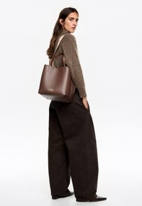 Bolso tote de cuero marrón con una textura suave, forma afilada y asas en contraste de color crema. Logo grabado en el frente. Diseñado con un estilo minimalista.