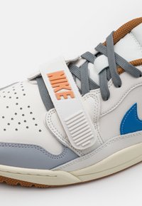 Gros plan d'une basket Nike blanche avec des lacets gris, un swoosh bleu, un embout perforé et une bande blanche étiquetée "NIKE" en orange.
