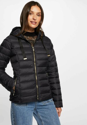 Chaqueta de plumas negra con capucha, diseño acolchado, cremallera frontal y acentos en dorado. Combinada con una camiseta negra y vaqueros azules.