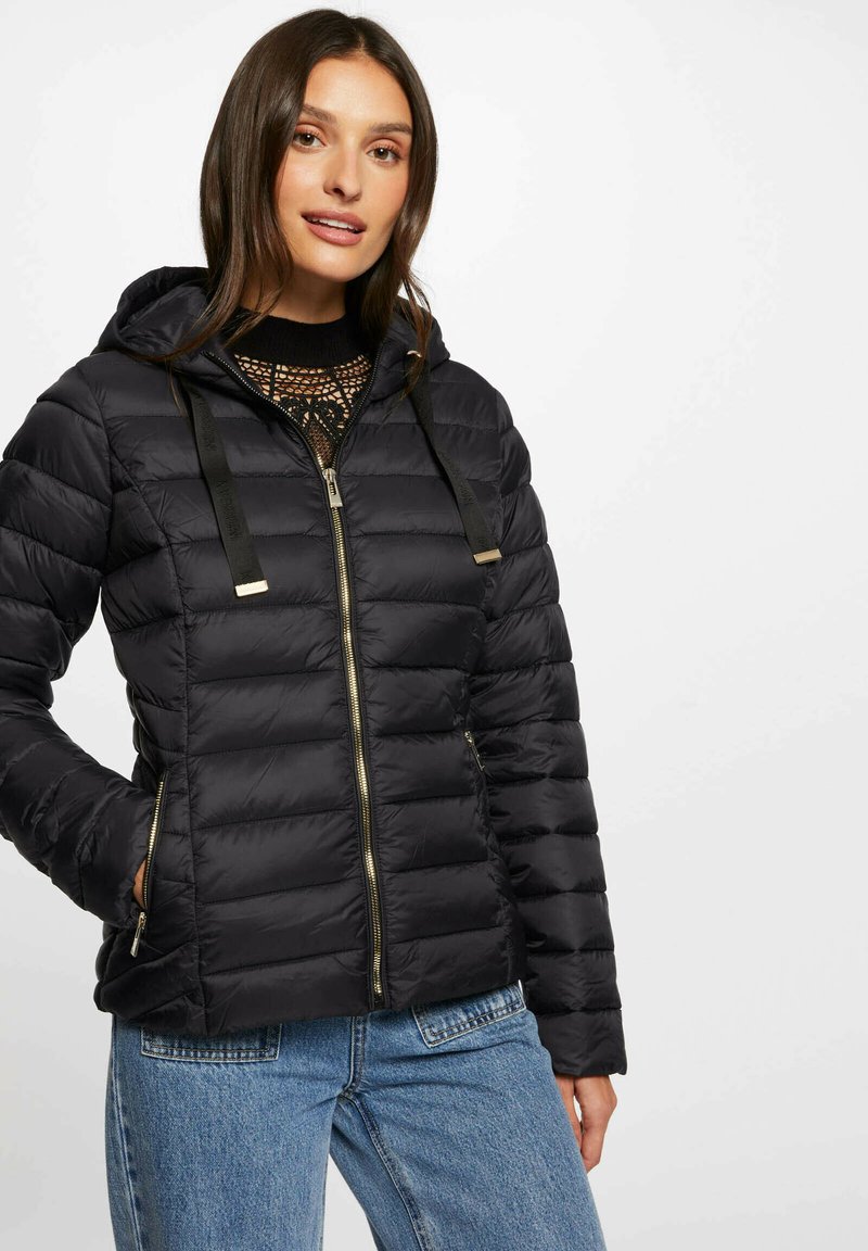 Giubbotto imbottito nero con cappuccio, design trapuntato, zip frontale e dettagli in metallo dorato. Abbinato a un top nero e jeans blu.