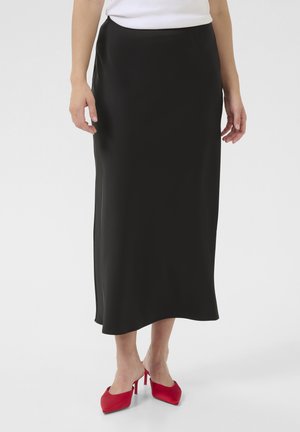 Černá saténová maxi sukně s hladkou texturou, s bočním rozparkem, nošená s červenými lodičkami na vysokém podpatku. Jednoduchý, elegantní design.