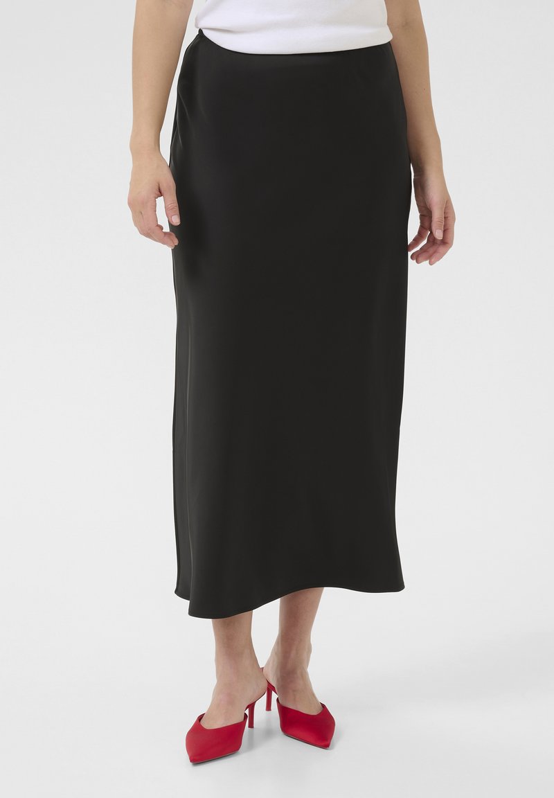 Čierna saténová maxi sukňa s hladkým povrchom, so bočným rozparkom, nosená s červenými vysokými opätkami. Jednoduchý, elegantný dizajn.